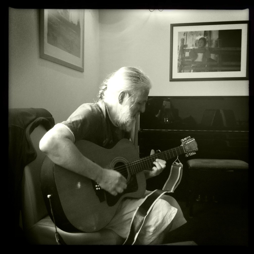 Monochrome picture of Andy Roberts playing an acoustic guitar unplugged in a small lounge bar with pictures on the wall