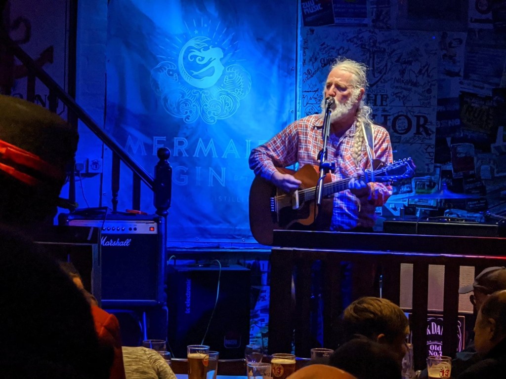 Andy Roberts playing guitar and singing into a microphone in a pub on the Isle of Wight 